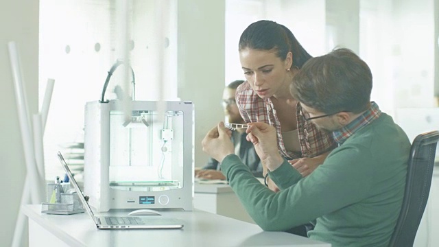 工程师使用3D打印机和讨论打印组件。他们的同事在后台工作。他们的位置是明亮的现代办公室。视频素材