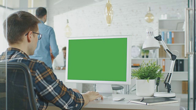 一个人坐在他的桌子上的绿色屏幕电脑的特写。背景模糊和明亮的办公室，人们通过办公室例行公事。视频素材