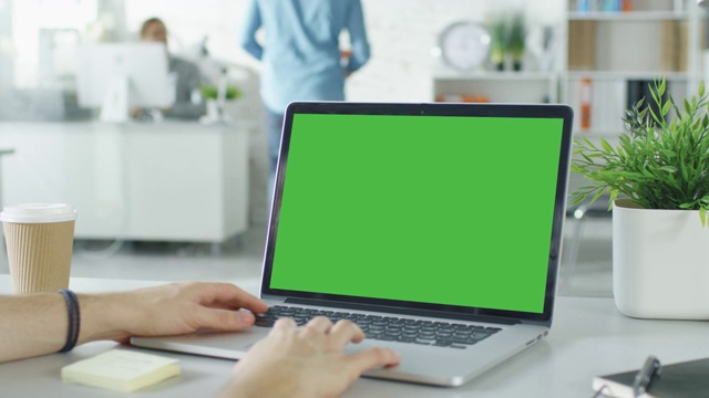一个男人的手在笔记本电脑的绿色屏幕上工作的特写。在背景模糊和明亮的办公室里，一个人靠近另一个人，他们在讨论。视频素材