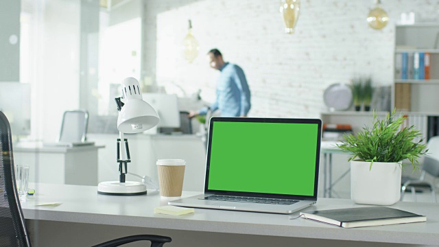 绿色屏幕的笔记本电脑站在桌子上，没有人在上面工作。背景里，一个男人走进来，坐在他的工作场所开始工作。视频素材