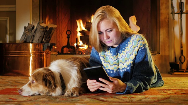 一个年轻漂亮的女人喜欢平板电脑。在一个以火为背景的舒适的房子里。它旁边是一只狗视频素材