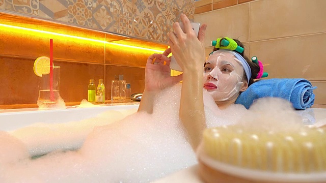 黑发女人戴着口罩在泡沫浴缸中使用手机。FullHD拍摄视频下载