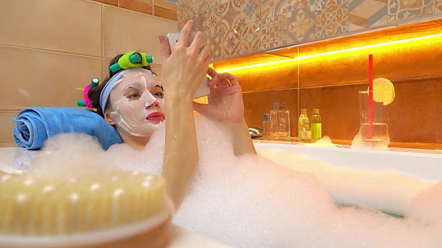 一名戴着面罩的黑发女子在充满泡沫的浴缸中轻拍智能手机。FullHD视频视频素材