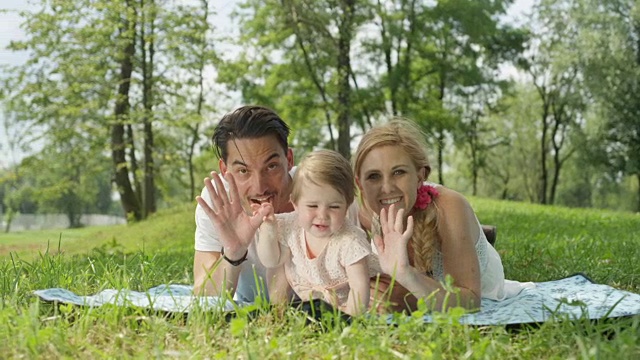 慢镜头:完美的年轻家庭和婴儿躺在毯子上在公园挥手视频素材