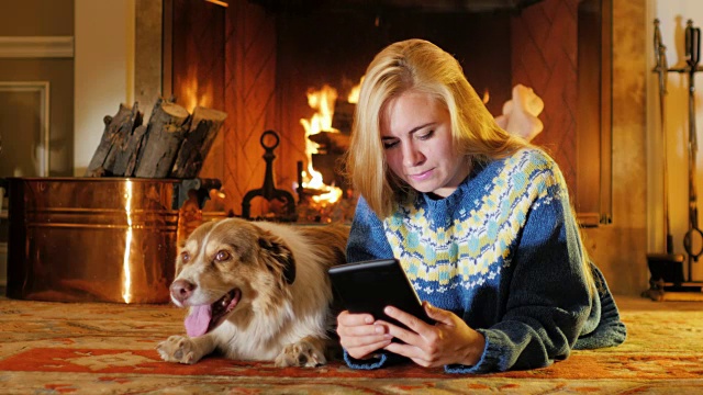 一个金发的年轻女子在燃烧的壁炉的背景下阅读电子书。它旁边是一条大狗视频素材