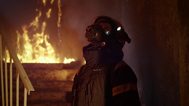 一个勇敢的消防队员检查燃烧的建筑物的肖像。他的手电筒打开了，他看着镜头。缓慢的运动。视频素材