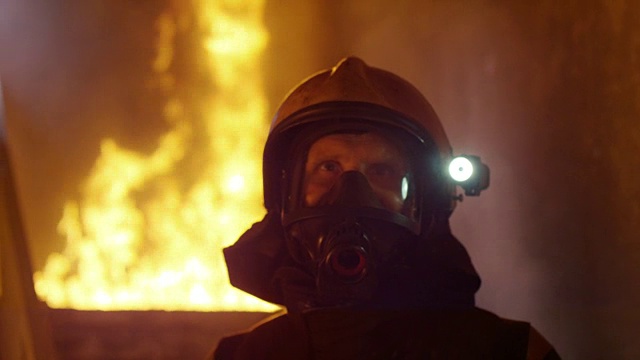 肖像拍摄的一个勇敢的消防员站在一个燃烧的建筑物熊熊大火在他身后。背景中的明火和烟雾。视频素材