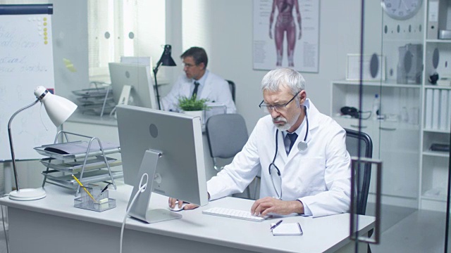 一个灰白头发的高级医生和他的助手在台式电脑工作的特写。坐在明亮的办公室里。视频素材
