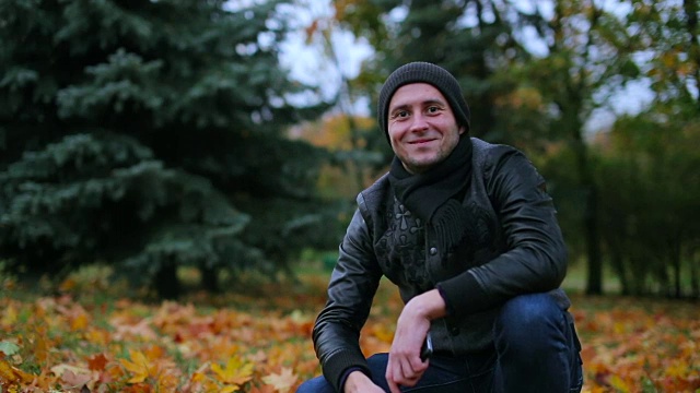 男人打招呼和挥手在相机里微笑坐在草地上的落叶背景-正面近距离肖像视频素材