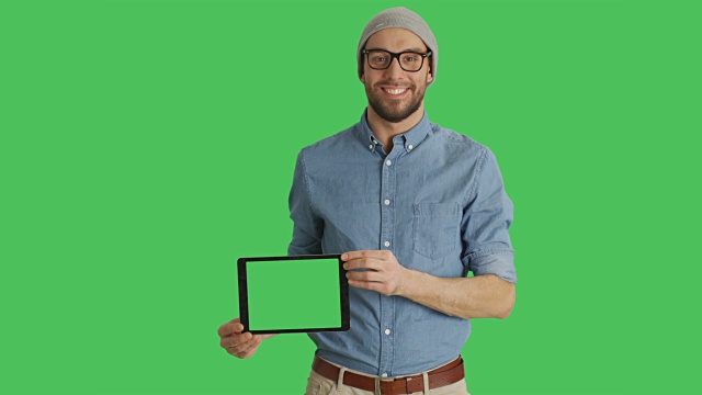 一个微笑的年轻人戴着眼镜展示绿色屏幕的平板电脑的中景。背景中的绿色屏幕。视频素材