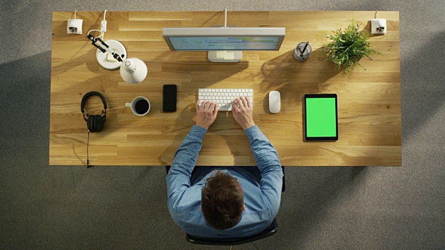 俯视图的软件工程师在他的桌面电脑坐在他的创意办公室。躺在桌上:智能手机，平板电脑，耳机。视频素材
