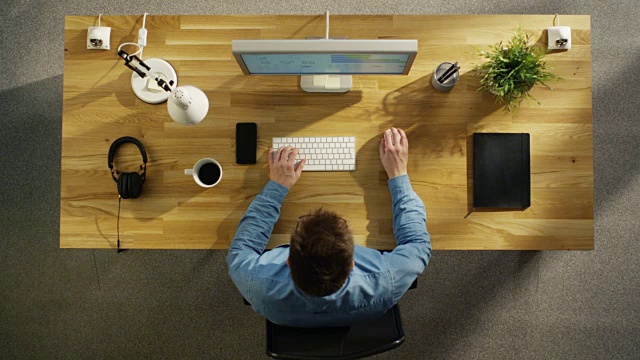 俯视图的一个软件工程师在台式电脑上工作，坐在他的桌子。桌子很整洁，智能手机，植物，咖啡杯，耳机都放在上面。视频素材