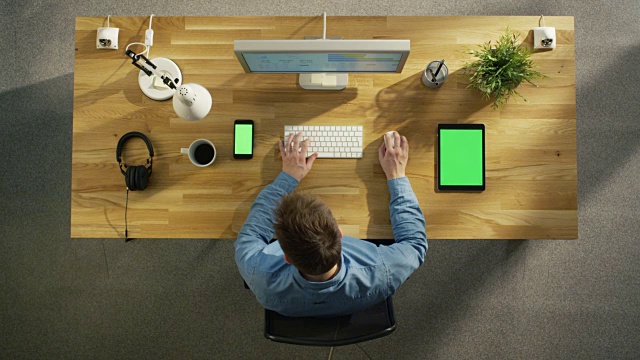 俯视图的软件工程师打字在他的桌面电脑。桌上放着绿色屏幕的平板电脑和智能手机。视频素材