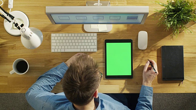 顶视图的年轻创意设计师看平板电脑与模拟绿色屏幕坐在他的办公桌。笔记本，个人电脑和咖啡杯也在桌子上。视频下载
