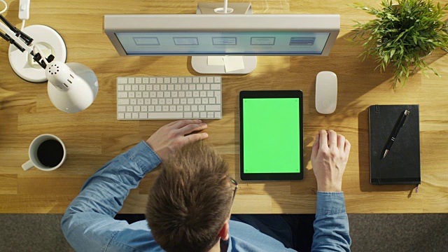 俯视图的年轻创意设计师轻敲平板电脑与模拟绿色屏幕坐在他的办公桌。笔记本，个人电脑和咖啡杯也在桌子上。视频素材