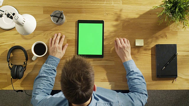 顶视图的年轻的创造性的人工作在他的绿色屏幕平板电脑坐在他的办公桌。他拿着他的笔和喝咖啡。视频素材