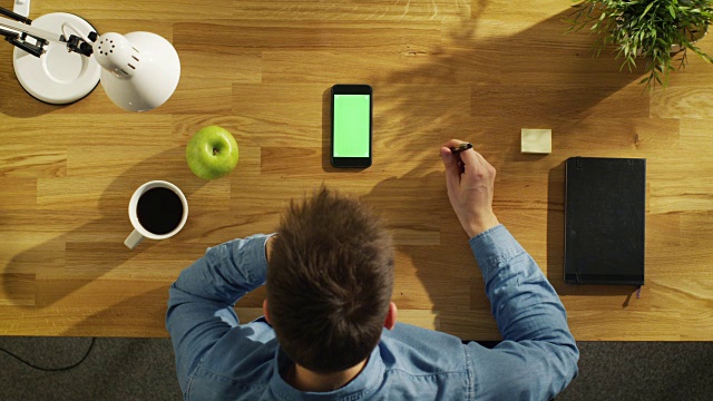 俯视图的年轻创造性的人在他的绿色屏幕智能手机上，而坐在他的办公桌。视频素材