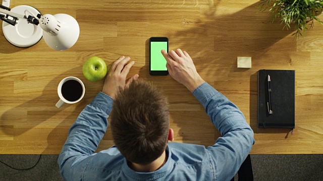 俯视图的年轻创造性的人在他的绿色屏幕智能手机上，而坐在他的办公桌。视频素材