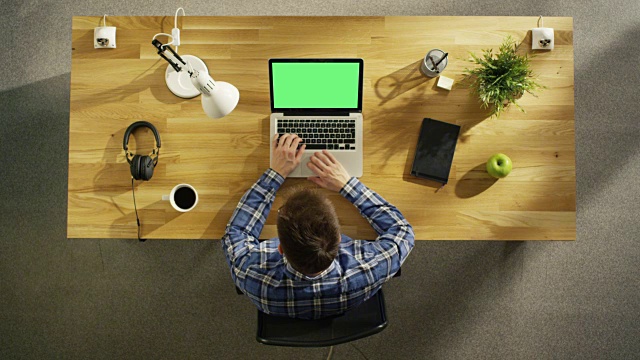 俯视图的一个年轻的创造性的人工作在他的绿色屏幕模拟笔记本电脑，而坐在他的木制书桌。桌子上也有:咖啡杯，智能手机，笔记本电脑，台灯，植物。视频素材