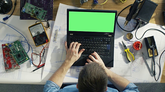 Top View of a Gifted IT技术员类型在他的绿色屏幕笔记本电脑上，而坐在他的办公桌，他被各种技术组件，草稿包围。阳光照在他的桌子上。视频素材