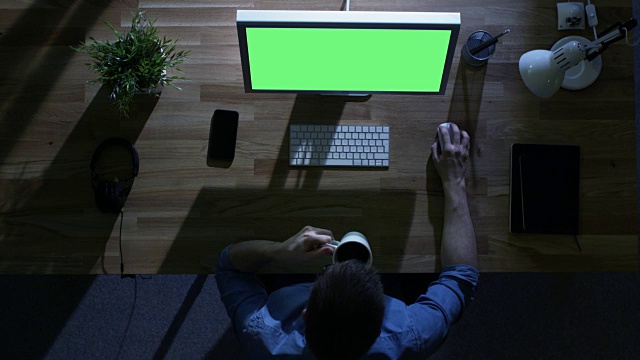 俯视图的男性程序员工作在他的桌面电脑与模型的绿色屏幕在晚上。他喝咖啡。他的桌子被外面的冷蓝光照亮。视频素材