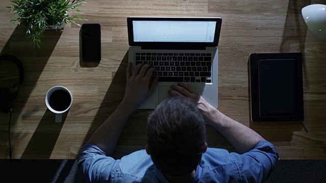 俯视图的创意设计师工作在他的笔记本电脑在晚上。他的桌子被外面的冷蓝光照亮。视频素材