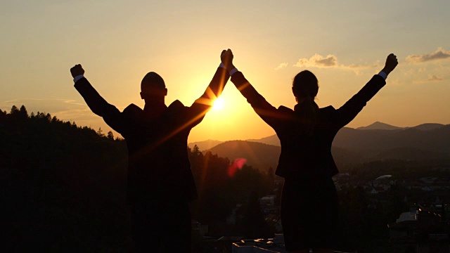 近景:日落时分，两名商人站在摩天大楼顶端，双手高举视频下载