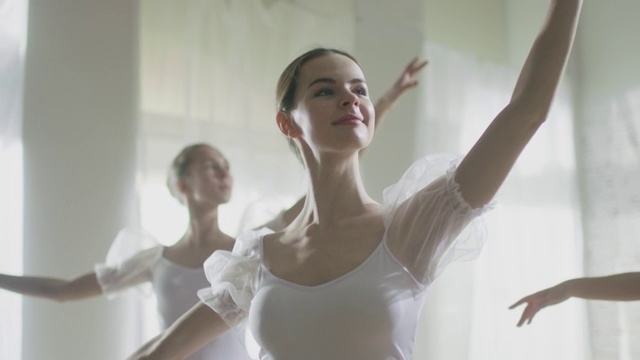 中景三个美丽和年轻的芭蕾舞者穿着白色芭蕾舞裙和跳舞。拍摄在一个阳光明媚的早晨在一个宽敞明亮的工作室。在缓慢的运动。视频素材
