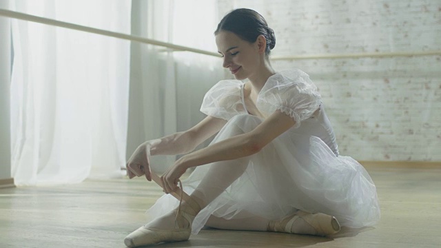 年轻美丽的芭蕾舞者坐在地板上她的芭蕾舞裙和系她的尖头鞋。在缓慢的运动。视频素材