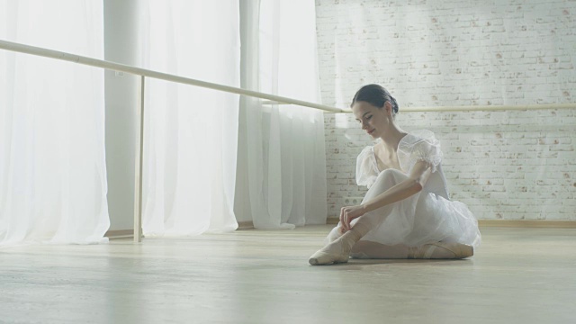 年轻美丽的芭蕾舞者坐在地板上她的芭蕾舞裙和系她的尖头鞋。视频素材