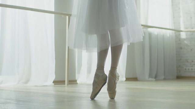 特写镜头的芭蕾舞女演员的腿。她穿着芭蕾舞鞋跳舞。她穿着白色芭蕾舞裙。在阳光明媚的现代工作室拍摄。在缓慢的运动。视频素材