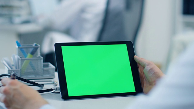 一个男医生使用绿色屏幕的平板电脑，他也拿着笔做笔记，他的助手在后台工作。办公室是明亮，白色和现代。视频素材