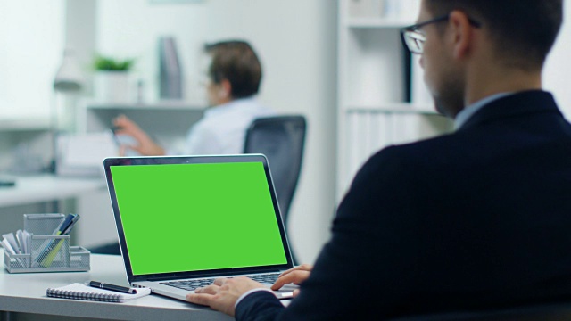 年轻的商人在绿屏笔记本电脑上工作。他的同事坐在办公桌前使用平板电脑。在现代灯光办公室拍摄。视频素材