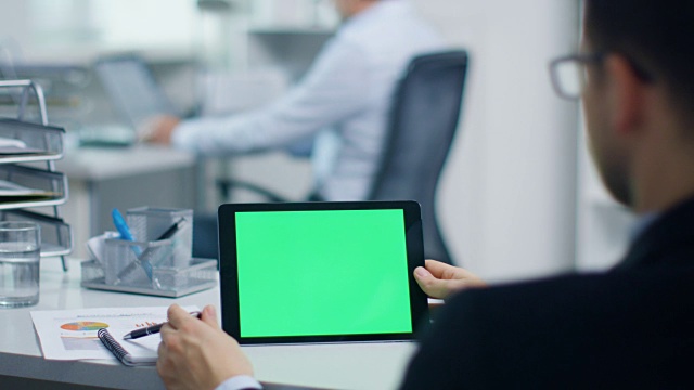 一个商人的特写工作在一个绿色屏幕的平板电脑上，握笔。他的同事在后台工作。办公室是光明和现代。视频素材
