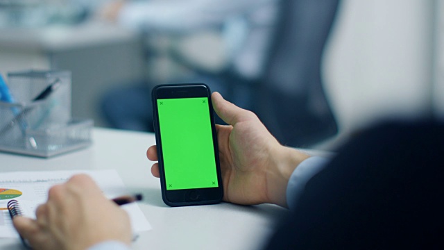 一个商人使用绿色屏幕的智能手机，在他的记事本上记笔记的特写。他的同事在笔记本电脑的后台工作。拍摄在一个明亮和现代的办公室。视频素材