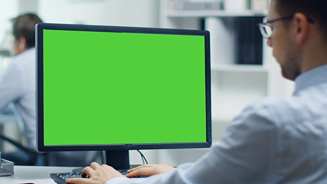 一个商人正在用绿屏的个人电脑工作。后台的同事。办公室是明亮和现代的。视频素材
