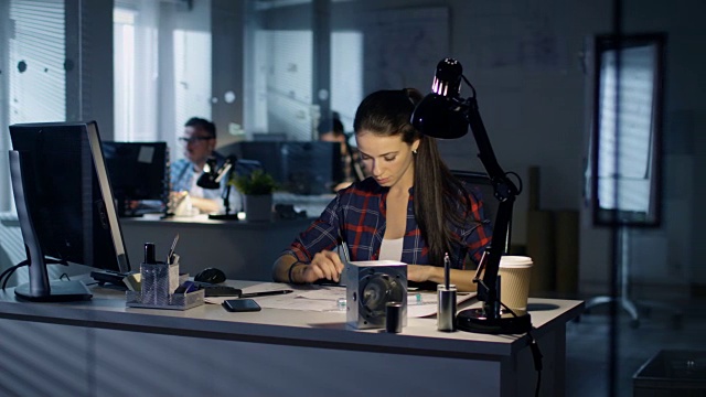 深夜，女性工业工程师坐在她的办公桌前起草方案。她的同事在后台工作。蓝色的城市之光透过窗户。视频素材