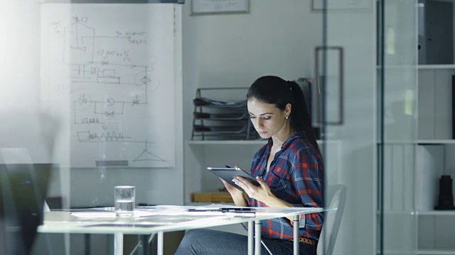 女设计工程师坐在办公室的玻璃桌前，用平板电脑工作，桌上放着设计图。的同事。在背景白板与图表和草图。视频素材