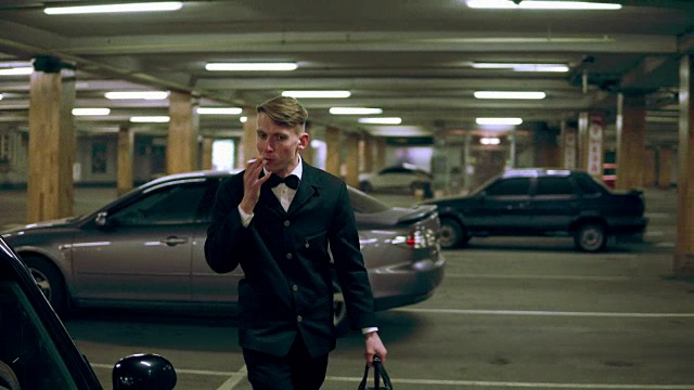 一个穿着黑色西装打着领结的年轻人抽着烟，停着车，晚上带着一个大黑包出去。视频下载