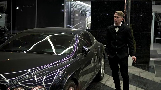 一个穿着黑色西装，打着领结的年轻人在夜晚的黑色豪华轿车旁抽着烟。视频下载