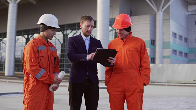 该项目的投资者身穿黑色西装，与身着橙色制服和头盔的建筑工人一起检查建筑对象。他们正在用石碑检查图纸。视频素材