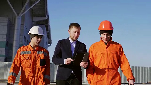 身穿西装的经理和两名身穿橙色制服、戴着头盔的工人正走过建筑设施。在4 k拍摄视频素材