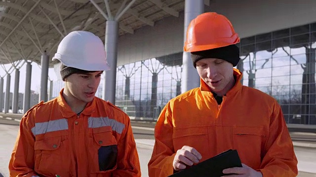 两名身穿橙色制服、头戴钢盔的建筑工人穿过建筑工地，一起查看设计图。背景建筑视频素材