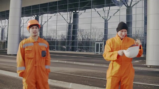 两名身穿橙色制服、头戴钢盔的建筑工人走到一起。其中一个丢了头盔。玩得开心视频素材