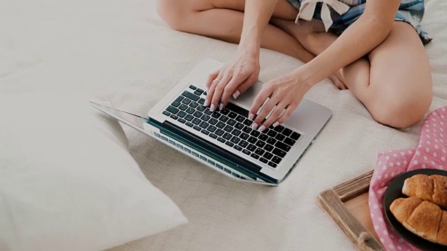 年轻女子坐在家里白色的床上使用无线技术。女孩在笔记本电脑上聊天的特写视频素材