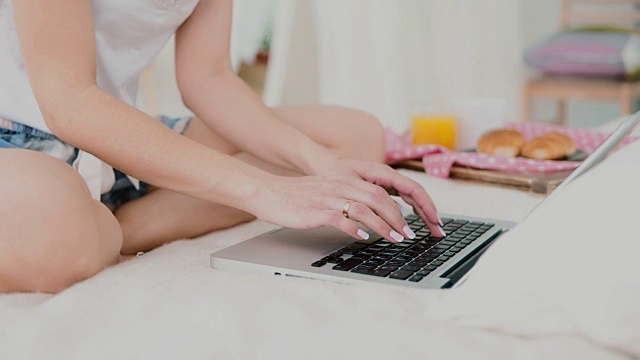 年轻女子坐在白色的床上用笔记本电脑吃早餐。女孩聊天和喝果汁的特写镜头视频素材