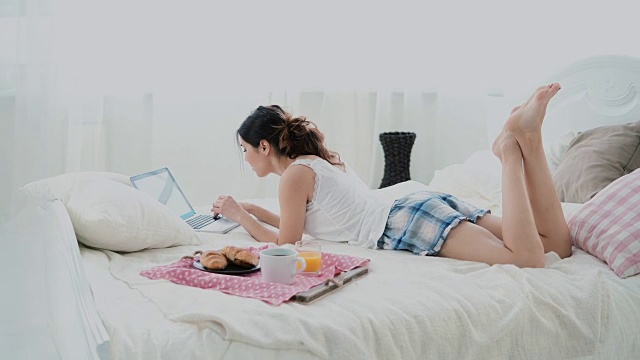年轻女子躺在家里的一张白色床上使用无线技术。吃早餐的时候用笔记本聊天的女孩视频素材