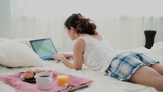 年轻女子躺在家里白色的床上用笔记本电脑吃早餐。黑发女孩在电脑上打字视频素材