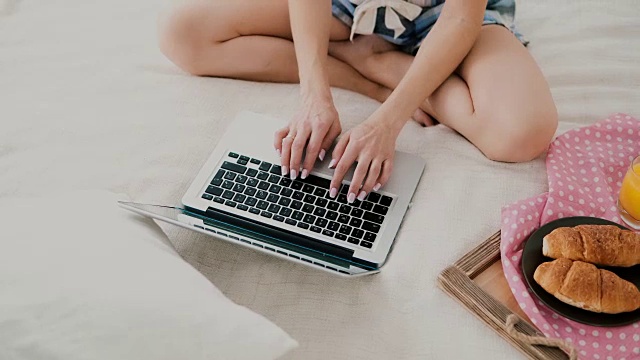 年轻女子坐在家里白色的床上用笔记本电脑吃早餐。特写的女孩打字电脑视频素材