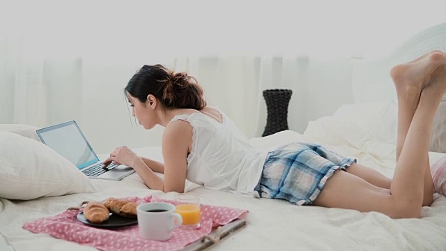 年轻女子躺在家里白色的床上用笔记本电脑吃早餐。黑发女孩一边在电脑上打字一边吃牛角面包视频素材